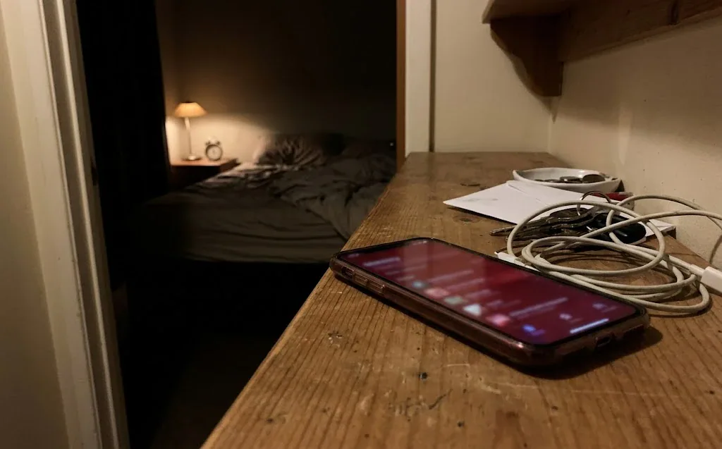 Smartphone op bureau met kabels, slaapkamer op achtergrond.